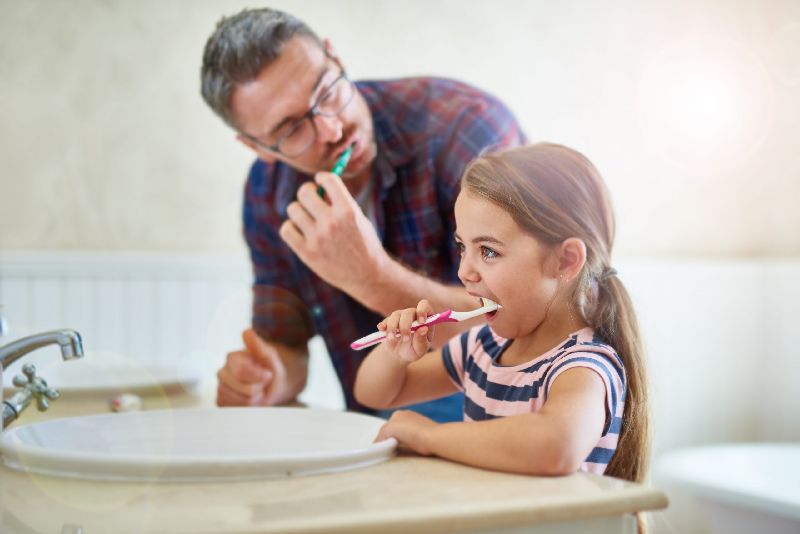 A father and daughter brushing their teeth together at a bathroom sink, promoting healthy dental habits.