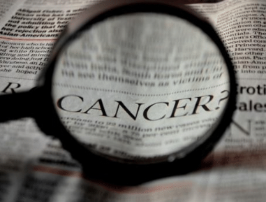 The word "CANCER" magnified through a glass lens, placed over a newspaper filled with text.