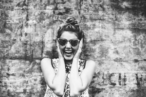 A woman smiling widely with her hands on her cheeks, wearing sunglasses against a textured wall background.