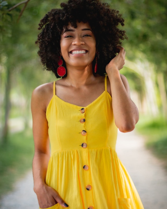 A smiling woman with curly hair wearing a yellow dress stands on a path lined with trees.
