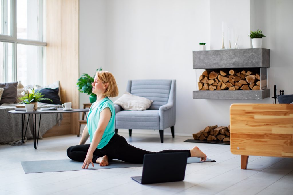 A woman in a turquoise shirt practices yoga on a mat in a bright, modern living room with a cozy fireplace.