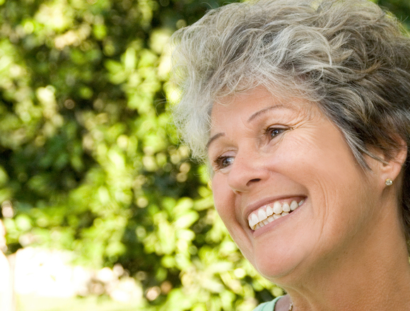 A smiling older woman with short, curly gray hair sits in front of lush green foliage.