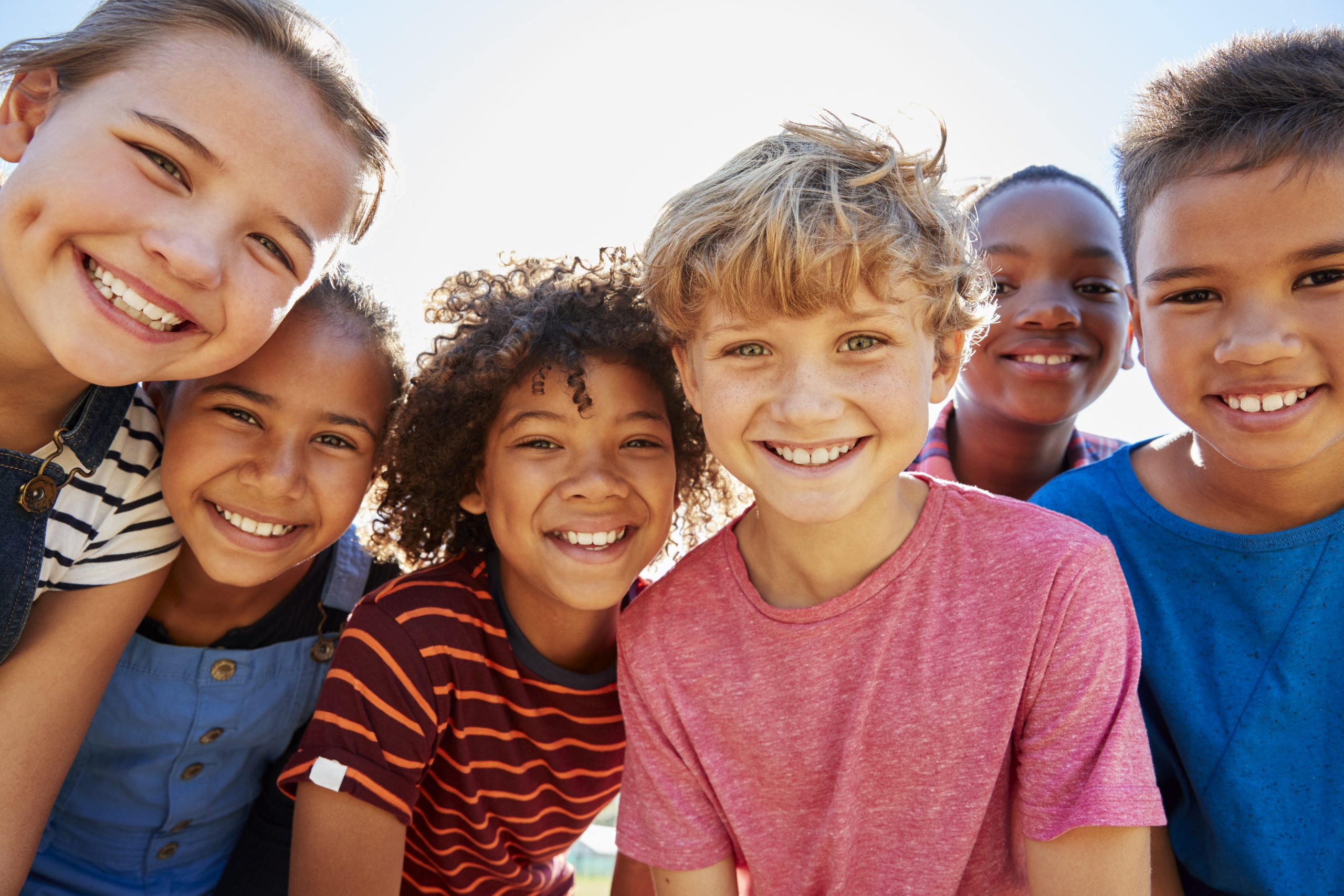 Six smiling children posing together outdoors, showcasing a variety of hairstyles and colorful clothing against a bright background.
