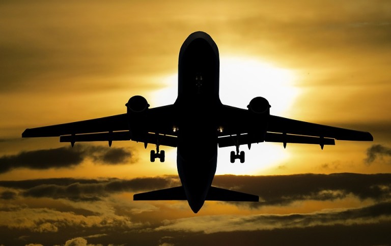 An airplane flying against a vibrant sunset with clouds, silhouetted against warm orange and yellow hues.