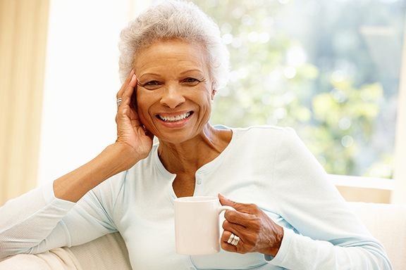 A smiling older woman holding a white mug sits comfortably on a sofa, surrounded by natural light.