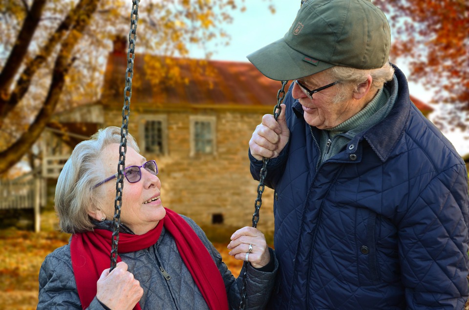 An elderly woman on a swing smiles at an elderly man in a cozy outdoor setting with autumn foliage.