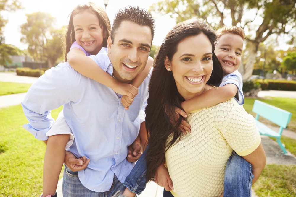 A smiling family of four enjoys a sunny day in a park, with two children playfully riding on their parents' backs.