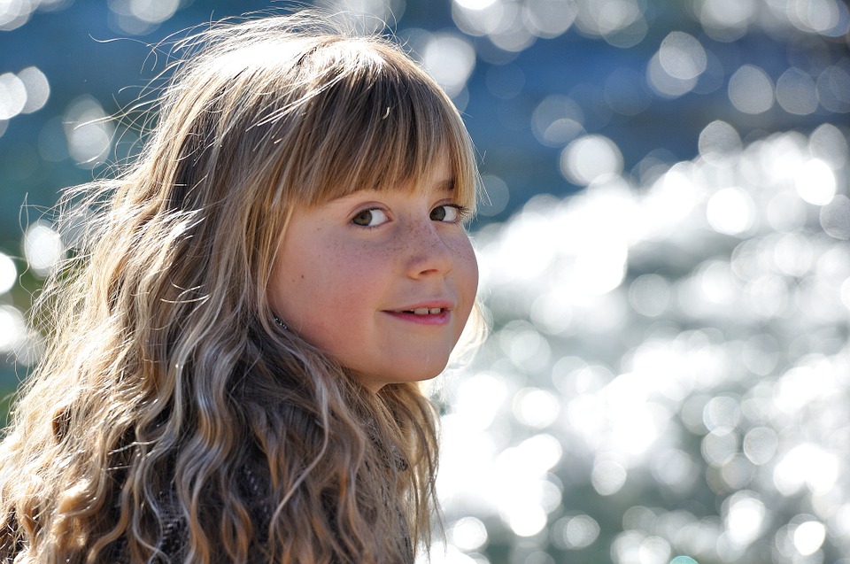 A young girl with long, wavy hair smiles while standing near shimmering water, with sunlight creating a bokeh effect in the background.