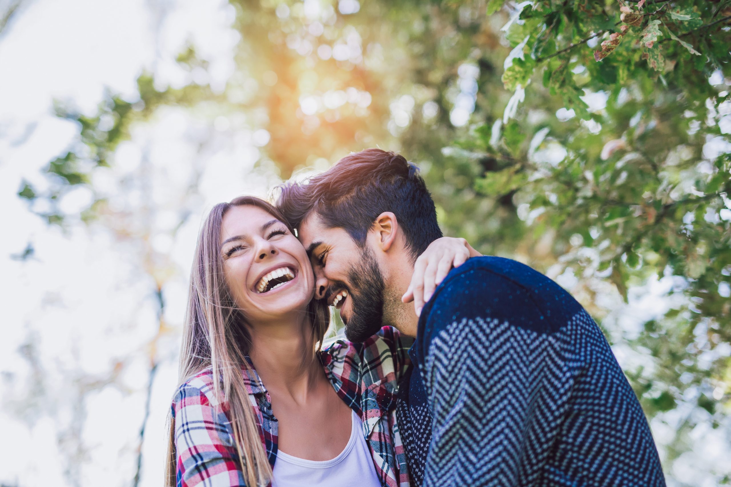 A young couple laughing together outdoors, surrounded by green trees and a bright, sunny atmosphere.