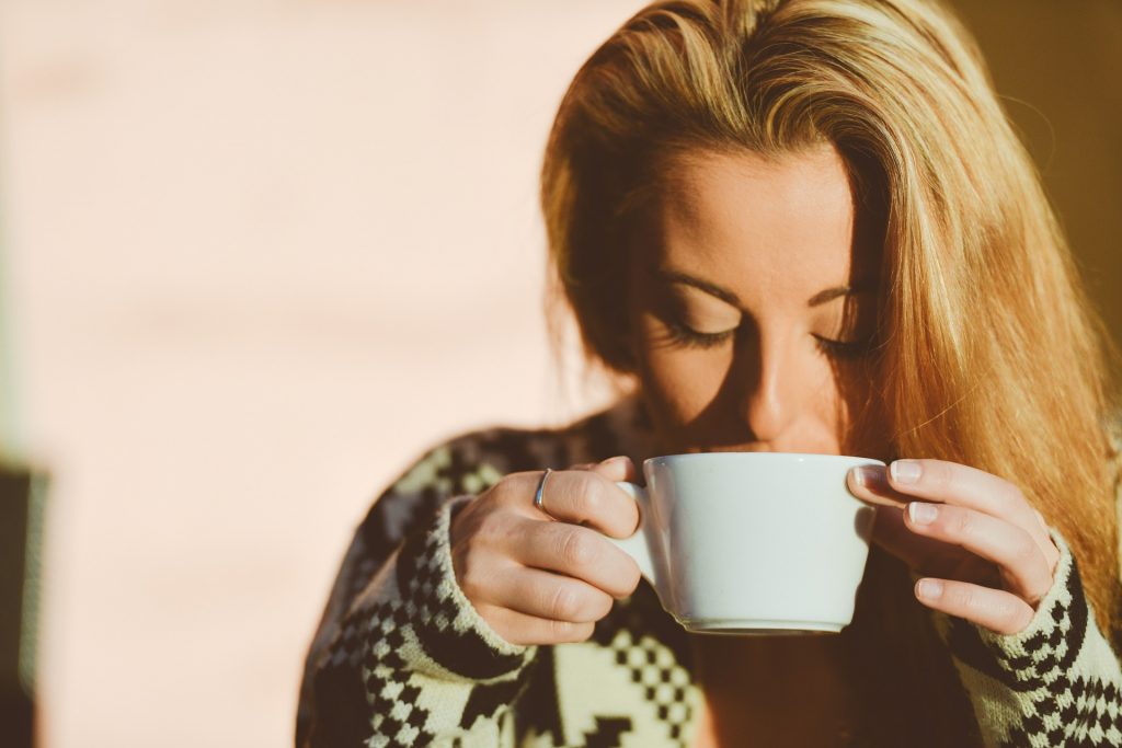 A woman with long blonde hair sips from a white cup while wearing a patterned sweater.