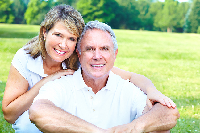 A smiling woman and man pose together outdoors, with the man seated and the woman's arms resting on his shoulders.