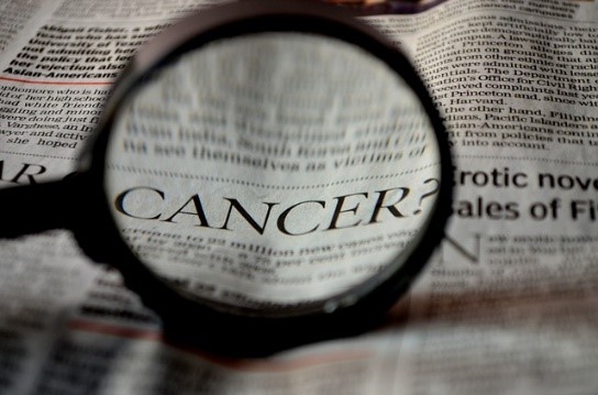 The word "CANCER?" is magnified through a lens on a newspaper page filled with text.