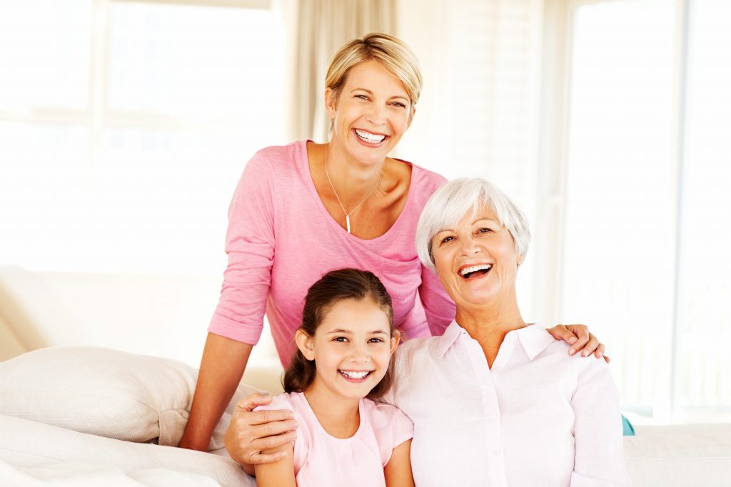 A smiling middle-aged woman stands behind a young girl and an older woman, all sitting together on a couch in a bright room.