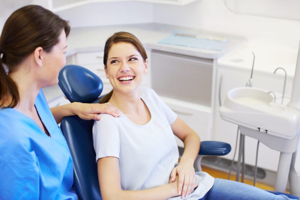 A patient smiling while sitting in a dental chair with a dentist's hand on her shoulder in a treatment room.