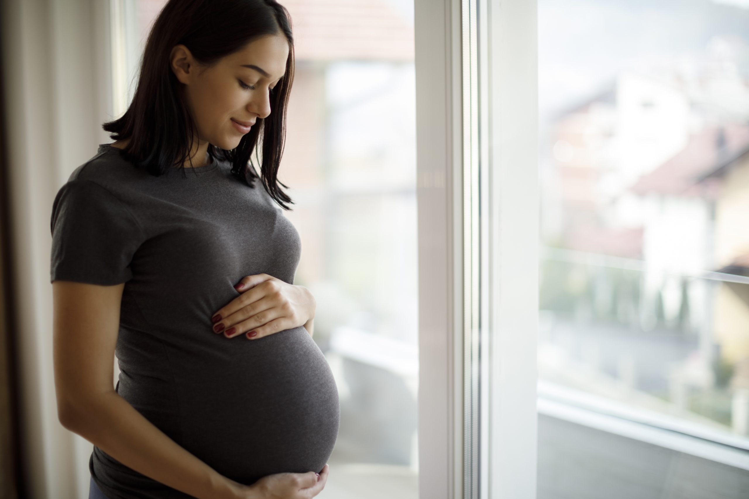 A pregnant woman gently cradles her belly while standing by a window in a bright interior space.
