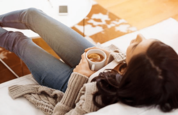 A woman relaxing on a couch, holding a cup of tea or coffee, with her legs crossed and a cozy sweater on.