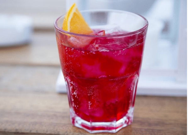 A glass of red soda with ice and a slice of orange garnish on a wooden table.