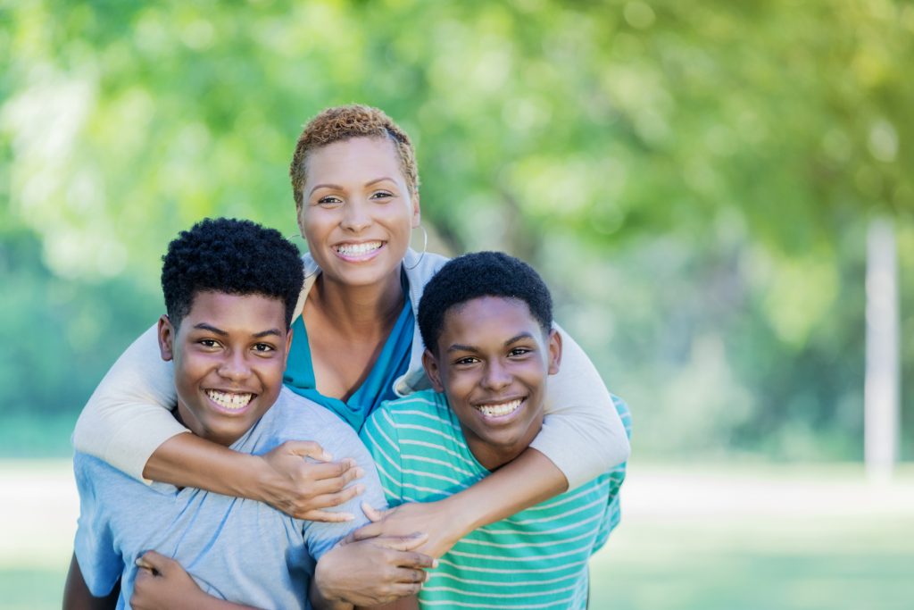 A smiling mother and her two sons pose together outdoors, with the mother embracing one son who is sitting on the other's back.