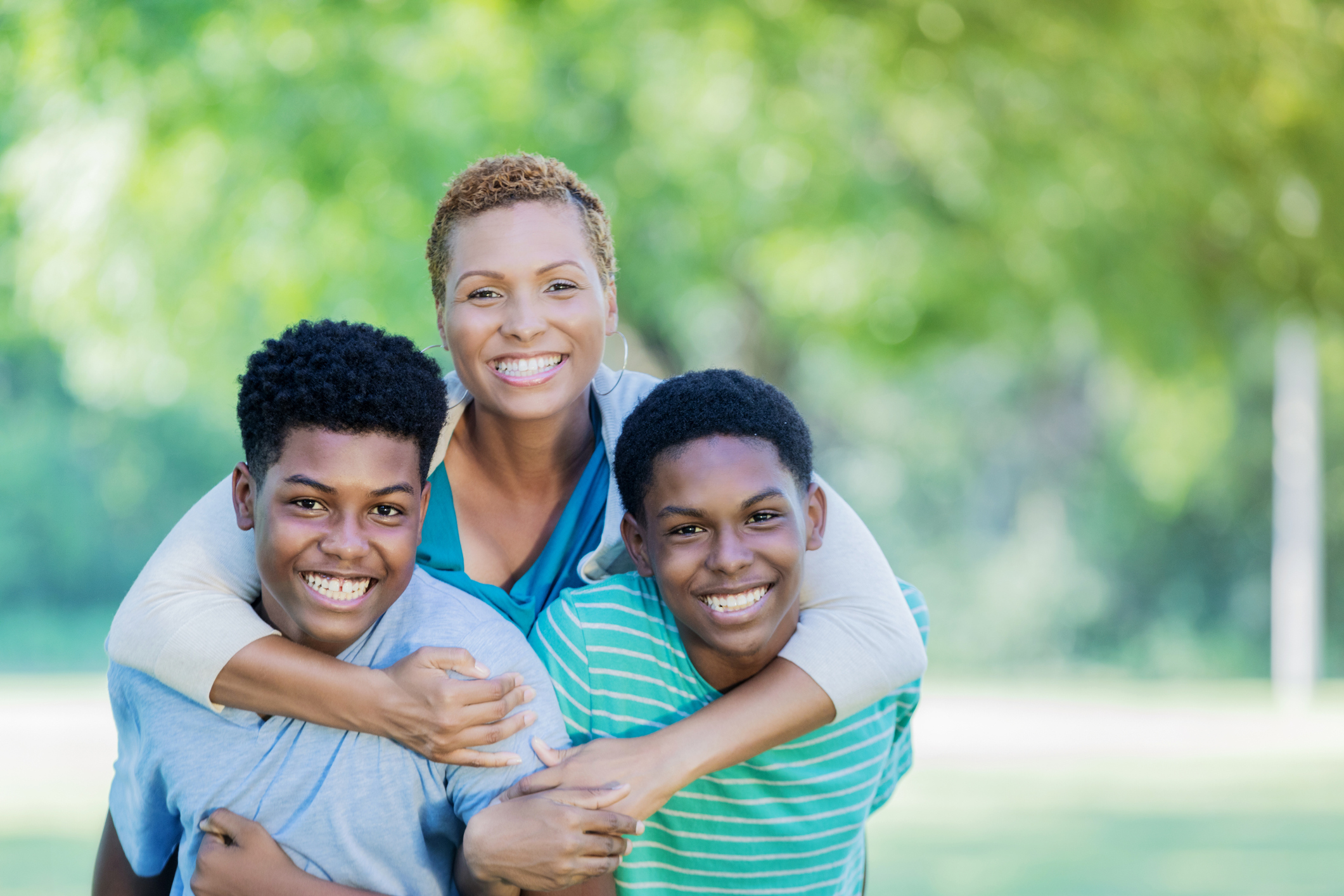A smiling mother and her two sons pose together outdoors, with the mother embracing one son who is sitting on the other's back.