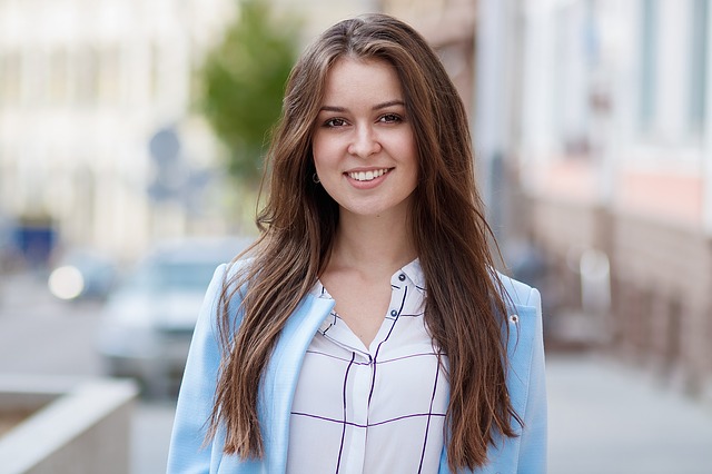 A woman with long brown hair smiles while wearing a light blue blazer and standing on a city sidewalk.