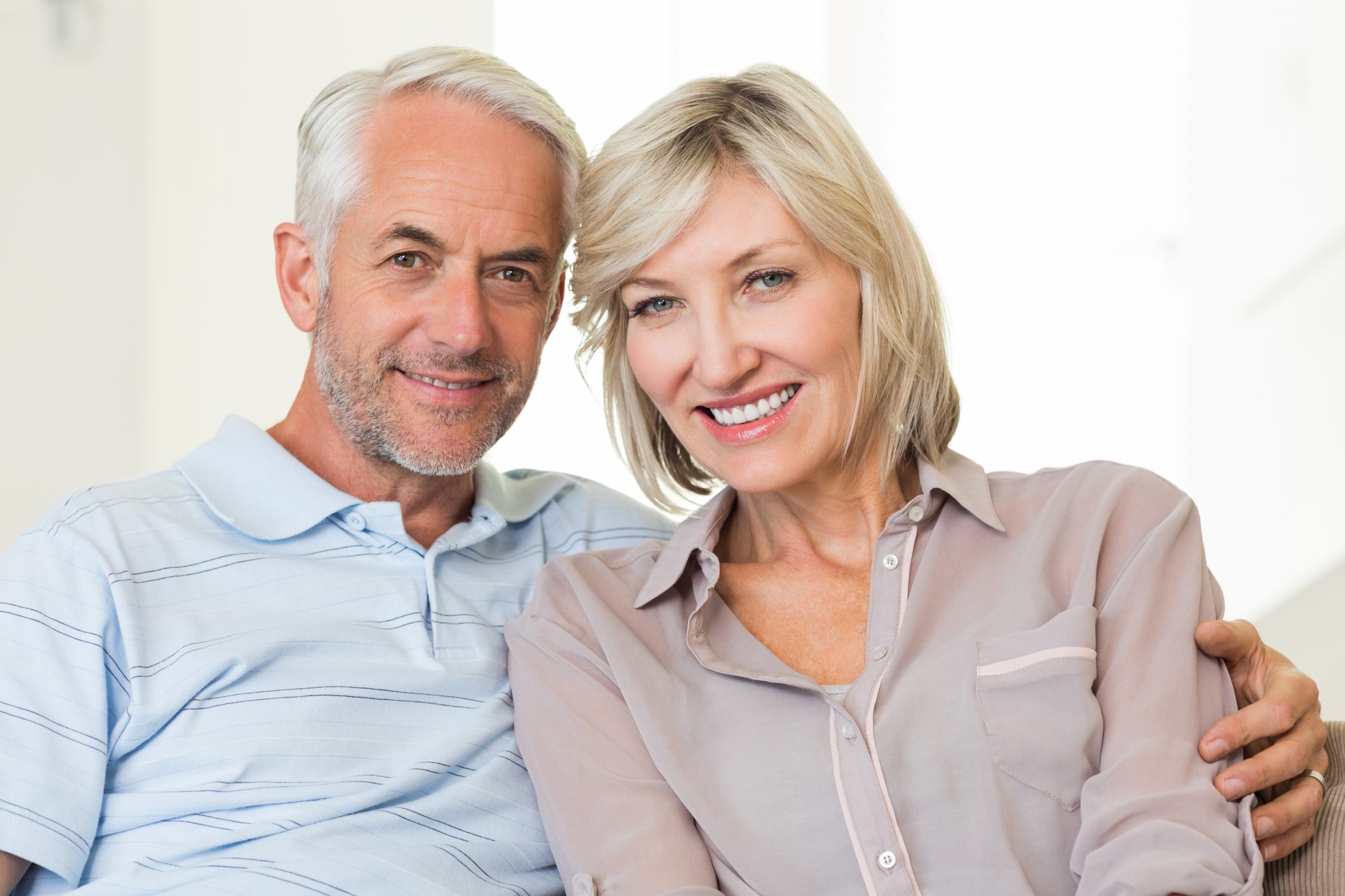 A smiling older couple sits together, showcasing their bright smiles in a well-lit indoor setting.