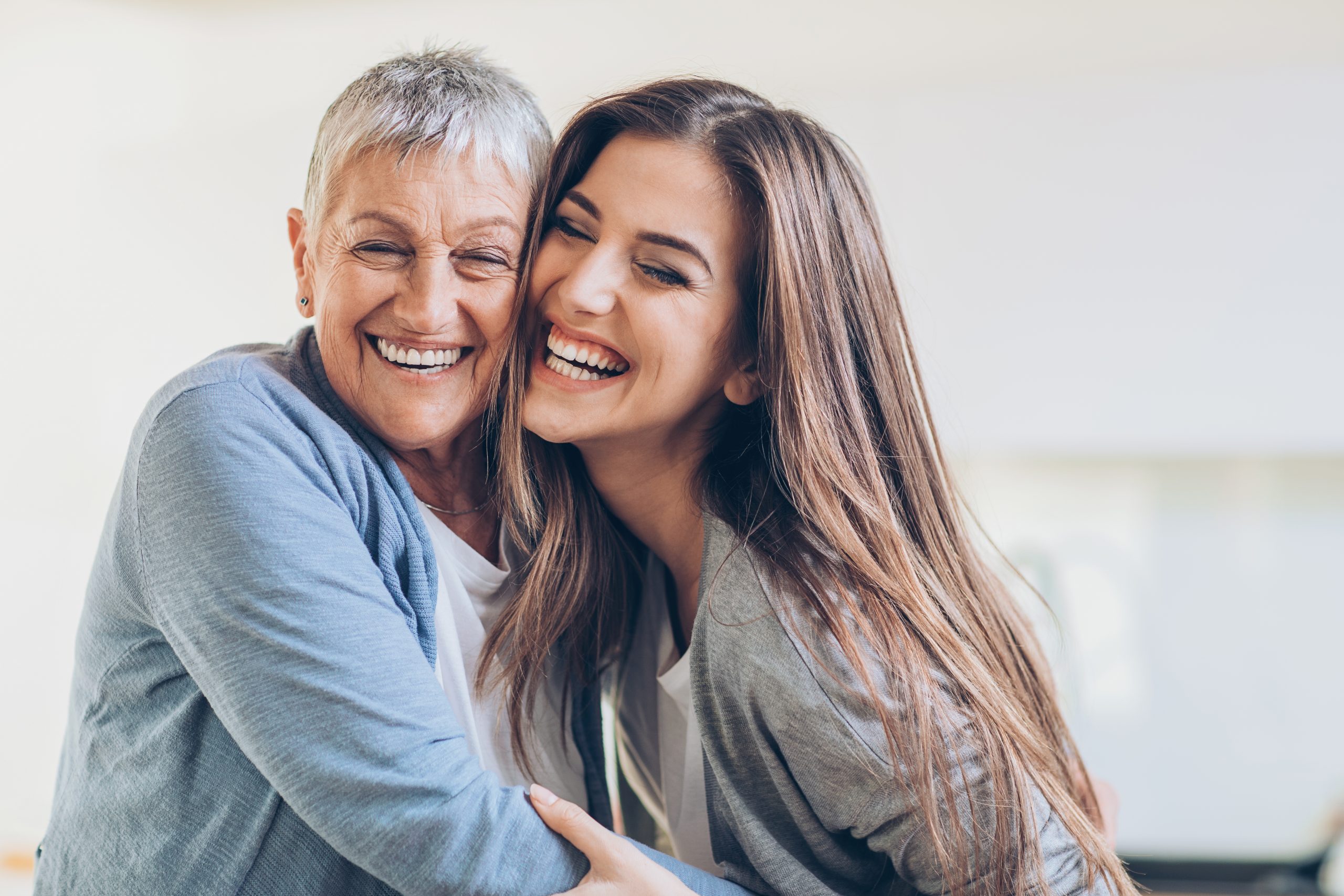 A smiling older woman and a young woman embrace joyfully, showcasing bright smiles in a bright, soft-lit room.
