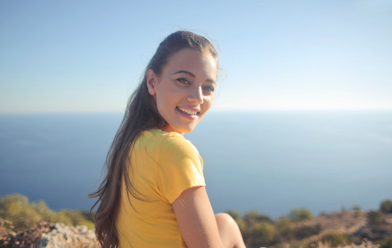 A woman with long hair smiles while sitting on a rocky hillside overlooking a calm blue ocean.