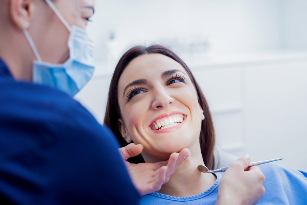 A dentist examining a smiling woman's teeth while she reclines in a dental treatment room.