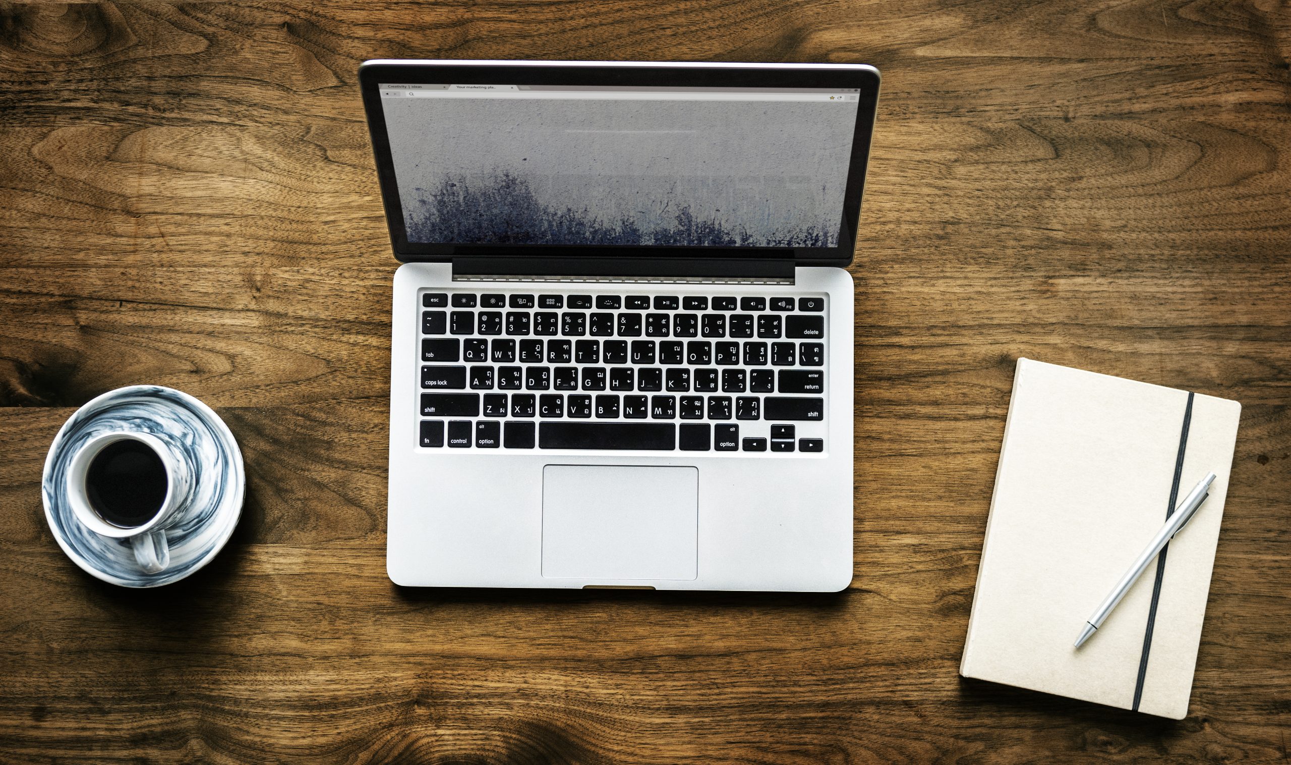 A laptop, a cup of coffee on a saucer, and a notebook with a pen are arranged on a wooden table.