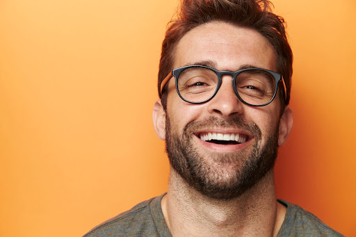 A smiling man with glasses and a beard against an orange background, showcasing healthy teeth, ideal for a dental promotion.