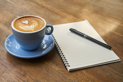 A blue coffee cup with latte art sits beside a blank spiral notebook and a pen on a wooden table.