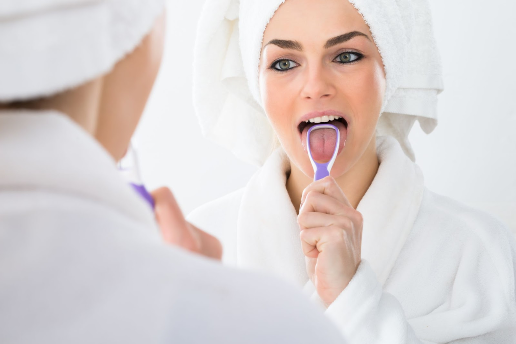 A woman in a white robe is using a tongue scraper while looking in a mirror.