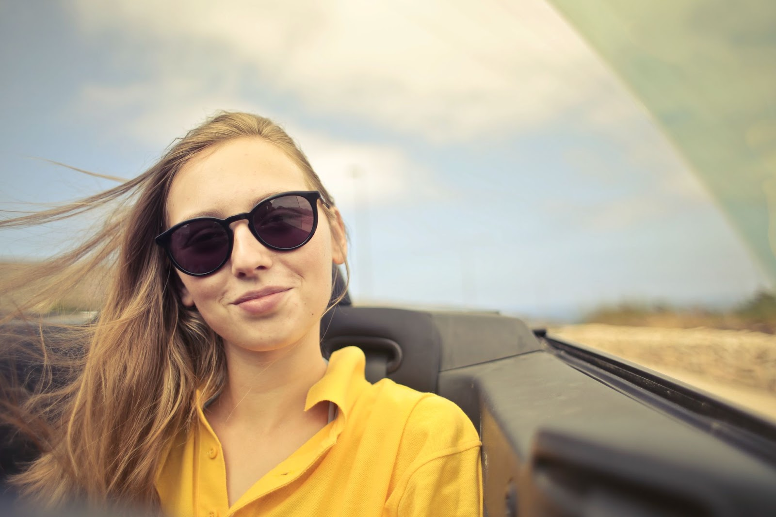 A young woman with long hair, wearing sunglasses and a yellow shirt, smiles in a convertible car under a bright sky.