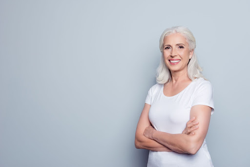 A smiling older woman with gray hair wearing a white shirt poses with crossed arms against a plain gray background.