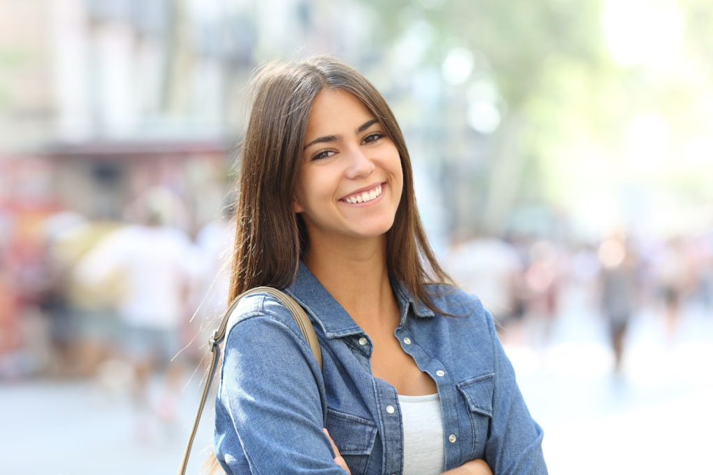 A young woman smiling in a busy street, wearing a denim jacket and a light top, with blurred pedestrians in the background.