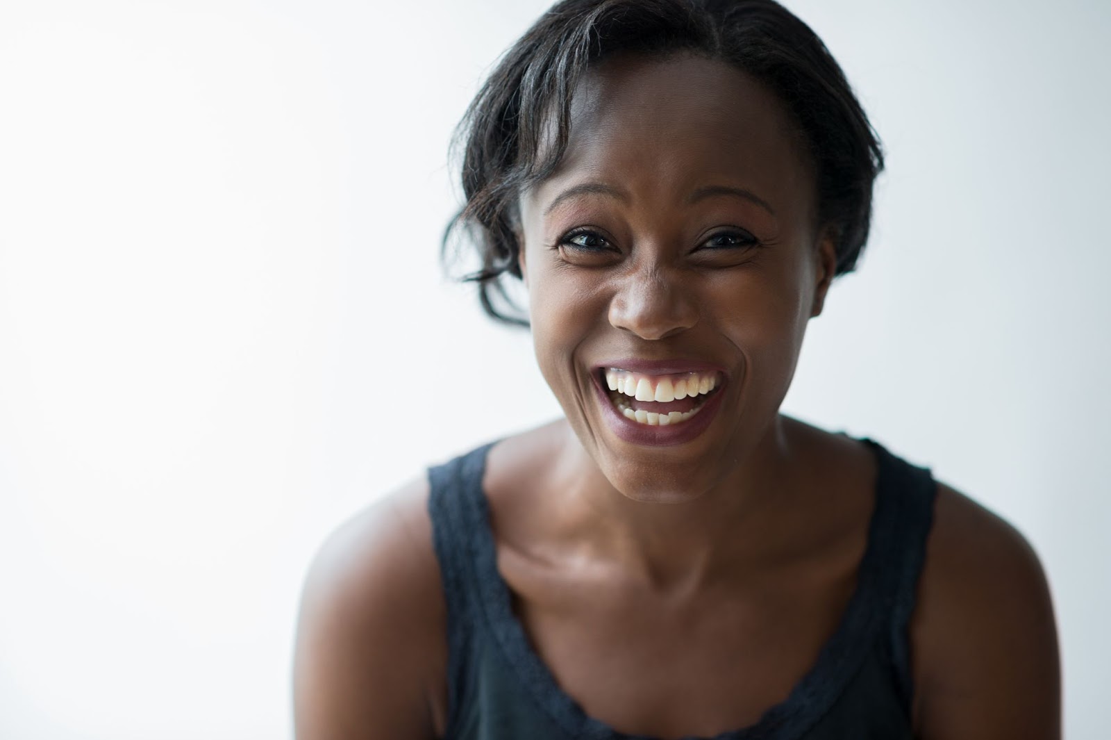 A woman with short hair smiles widely against a light background, showcasing her healthy teeth.
