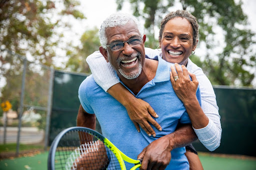 A smiling couple enjoys a playful moment on a tennis court, with the woman playfully hugging the man from behind.