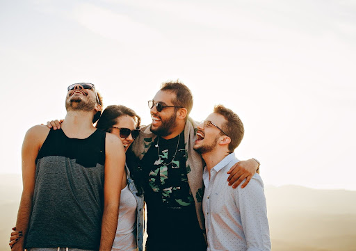 Four friends laughing and posing together outdoors against a bright sky, wearing sunglasses and casual clothing.