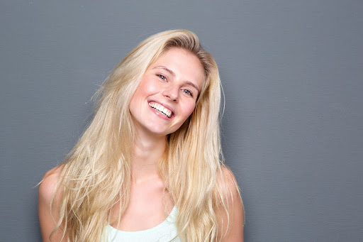 A young woman with long blonde hair smiling widely against a gray background.